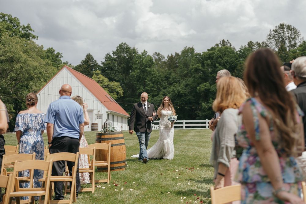 Wedding guests seated in wooden chairs on a lawn with a rustic barn visible in the background.