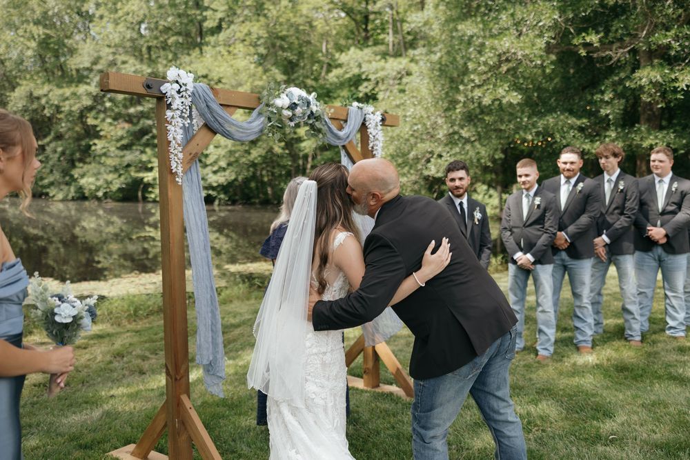 Wedding party plays a group game outdoors on a sloping green lawn surrounded by trees.
