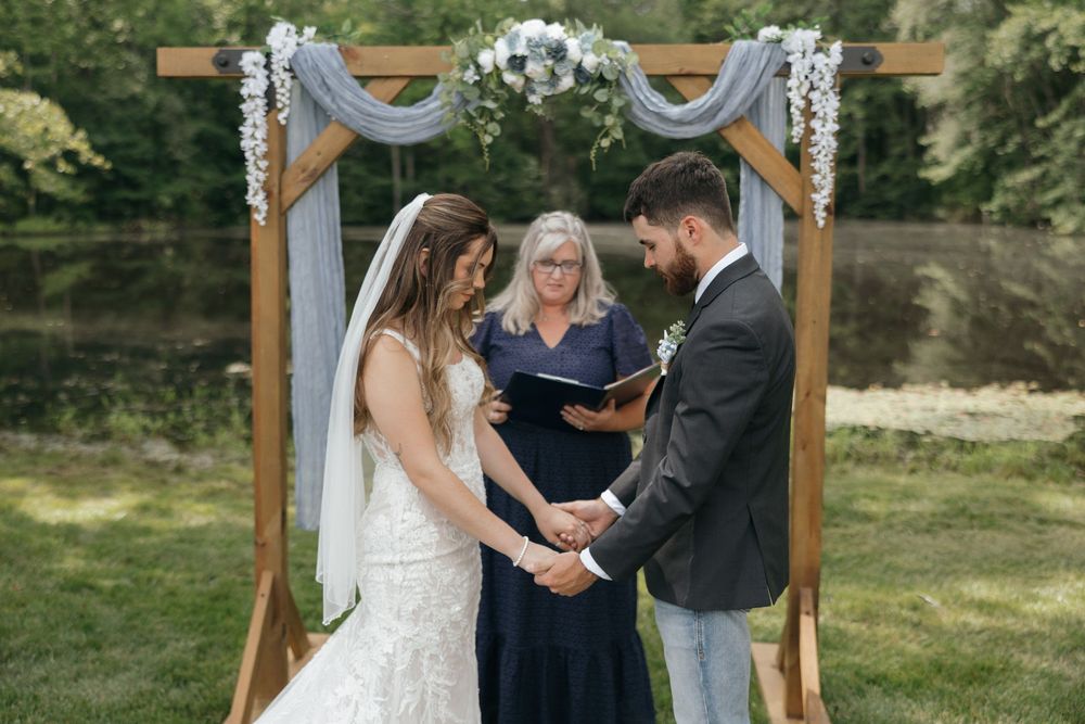 Couple exchanges rings during outdoor wedding ceremony beneath wooden arch decorated with white floral arrangements.