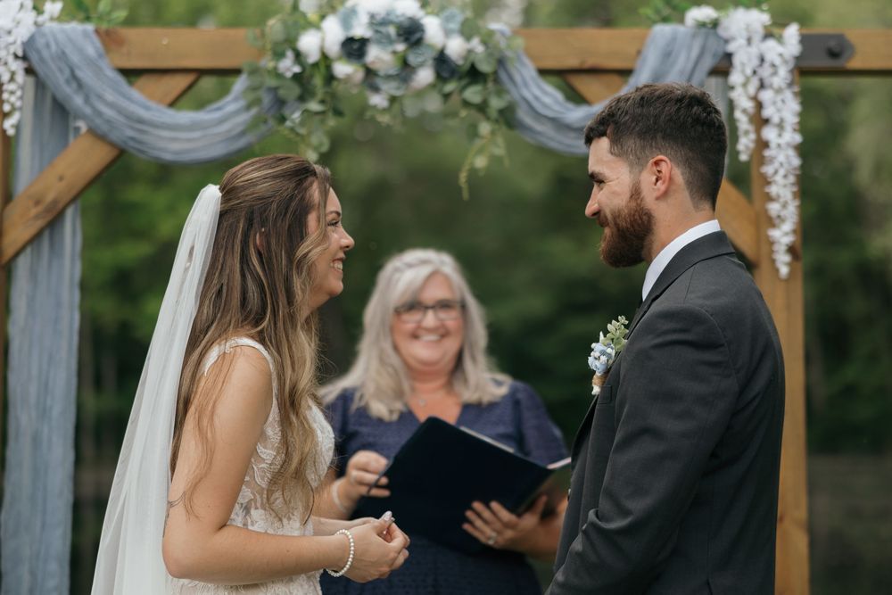 Wedding ceremony under a wooden arch decorated with white flowers and gray fabric in a garden setting.