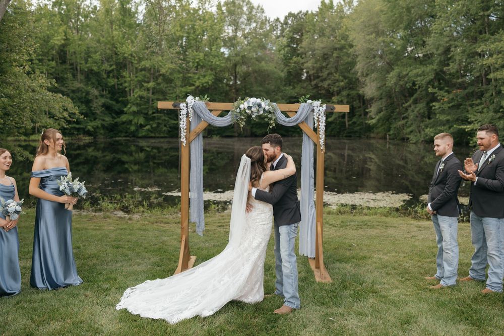 Outdoor wedding ceremony setup with wooden arch and guests seated on white chairs in a garden.
