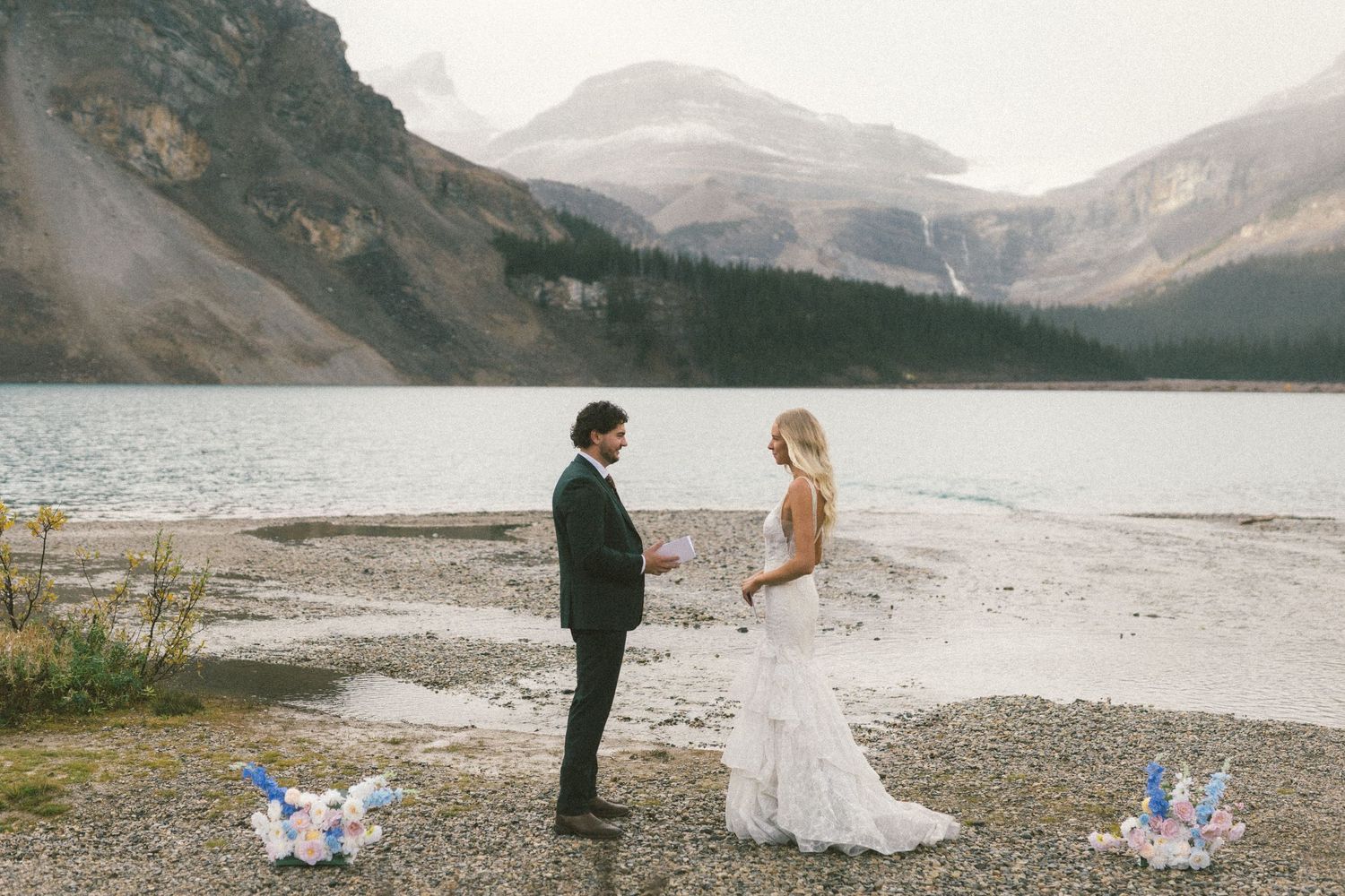bride and groom read each other vows on the shore of Bow Lake with mountains behind them