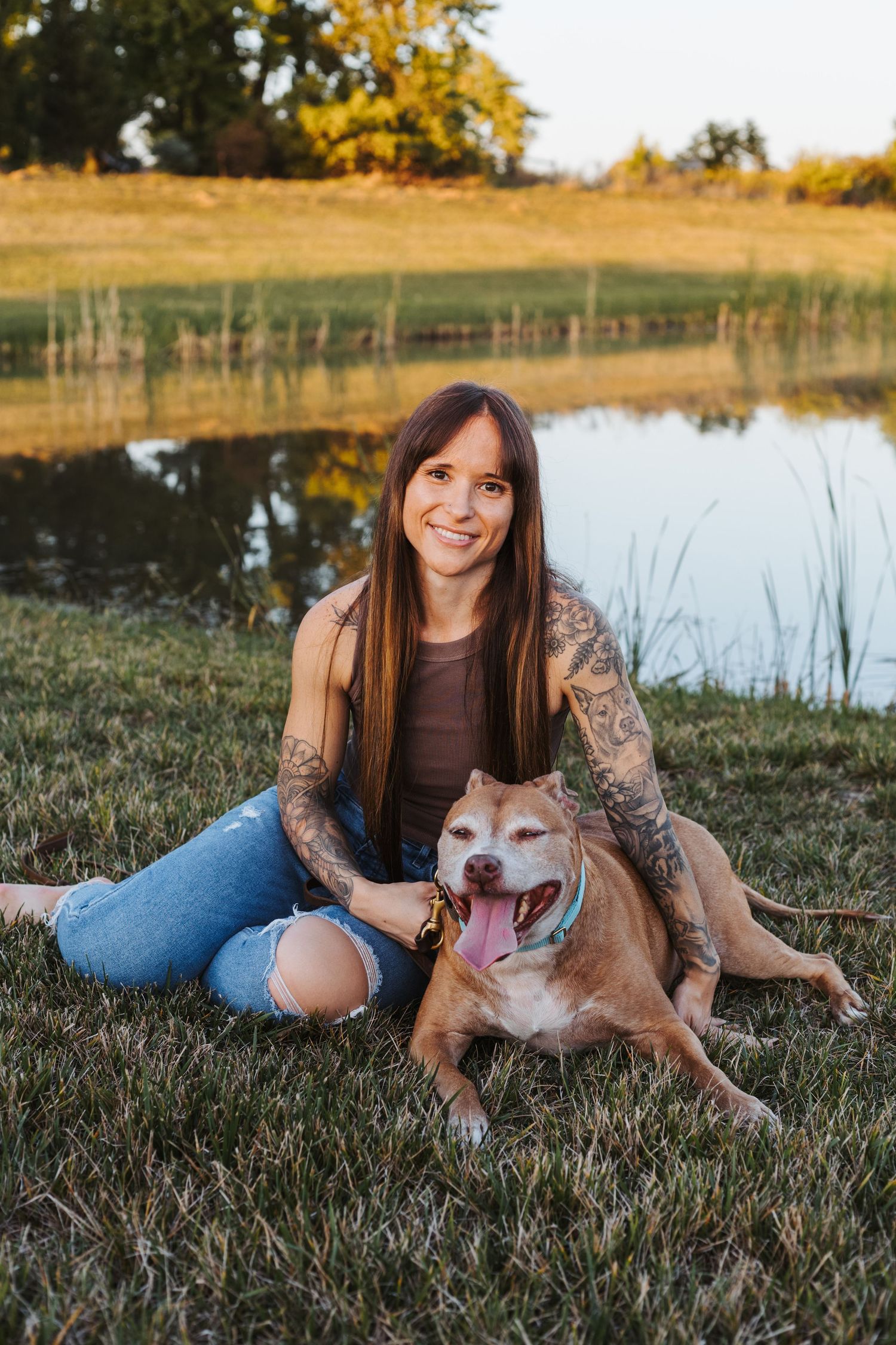A happy brown and white dog with a big smile lounges on grass by a pond during golden hour with its owner.