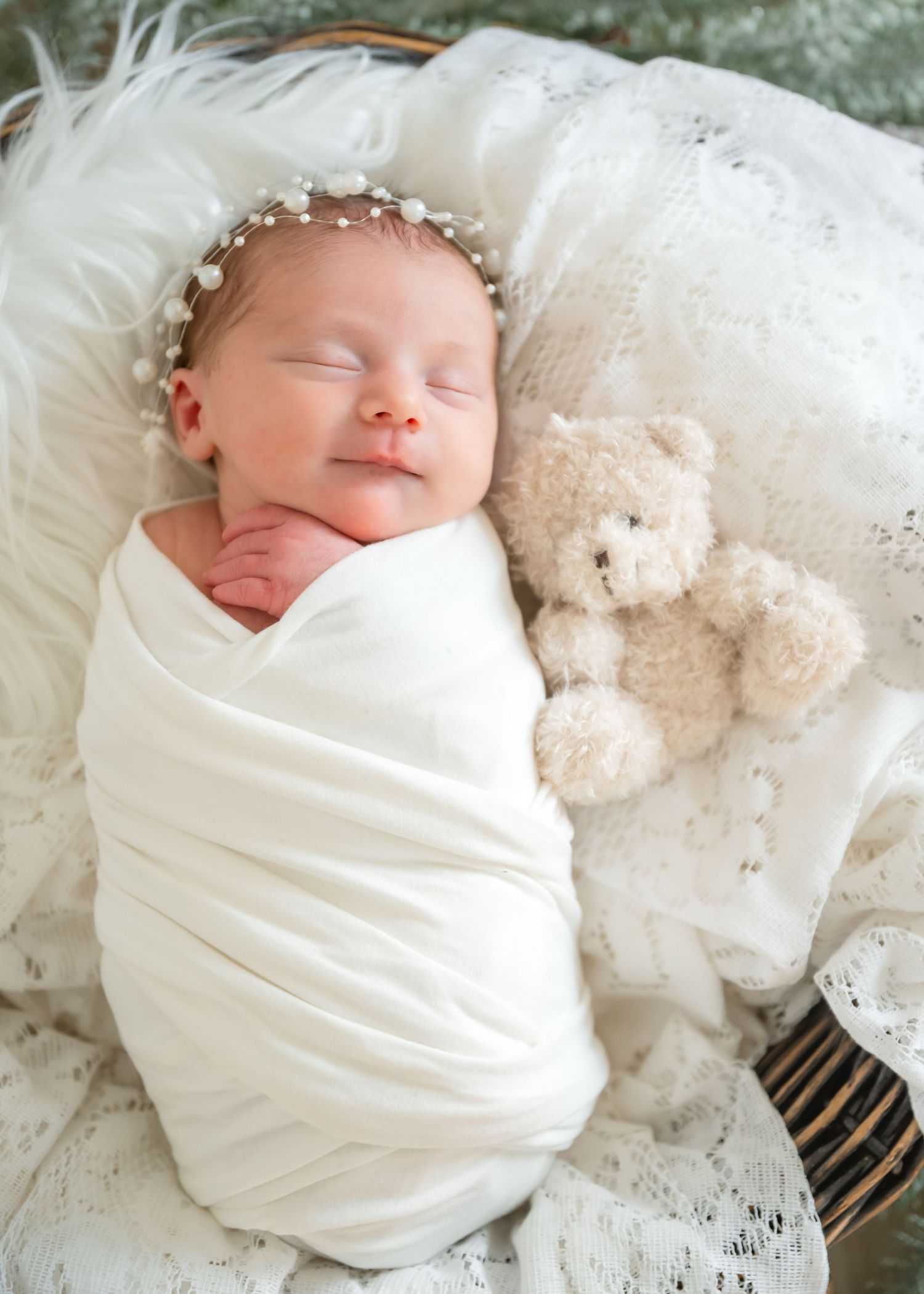 A sleeping newborn baby swaddled in white fabric lies peacefully next to a plush teddy bear on delicate lace bedding.