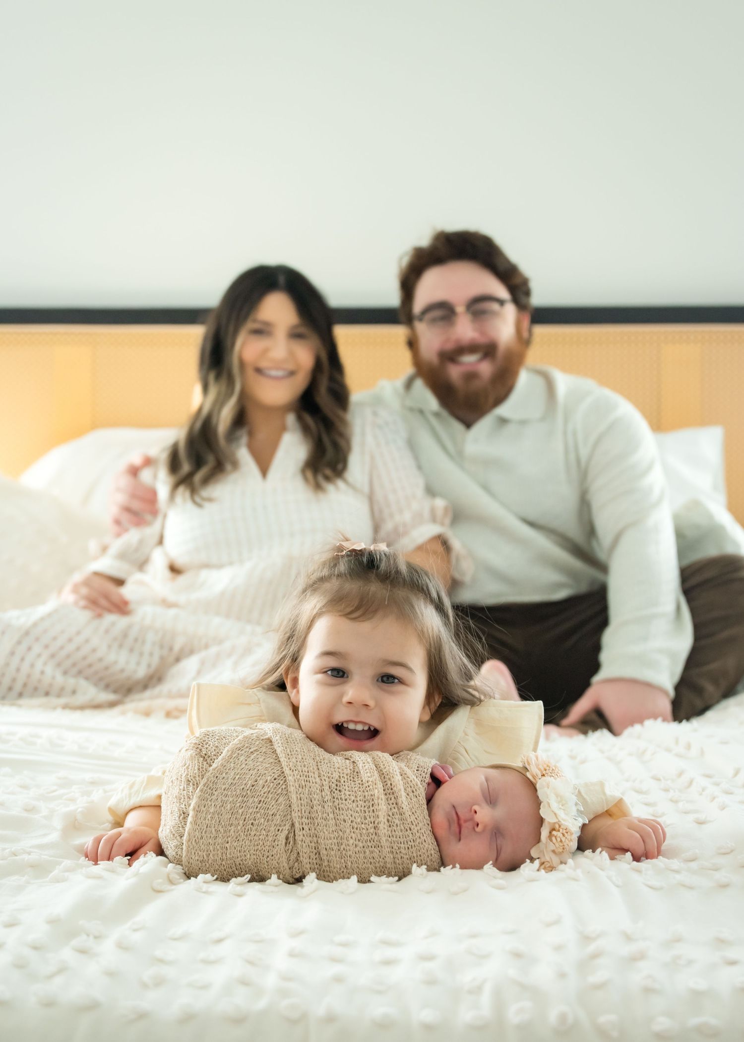 Family lounging together on white bedding while siblings share sweet moment with newborn baby.