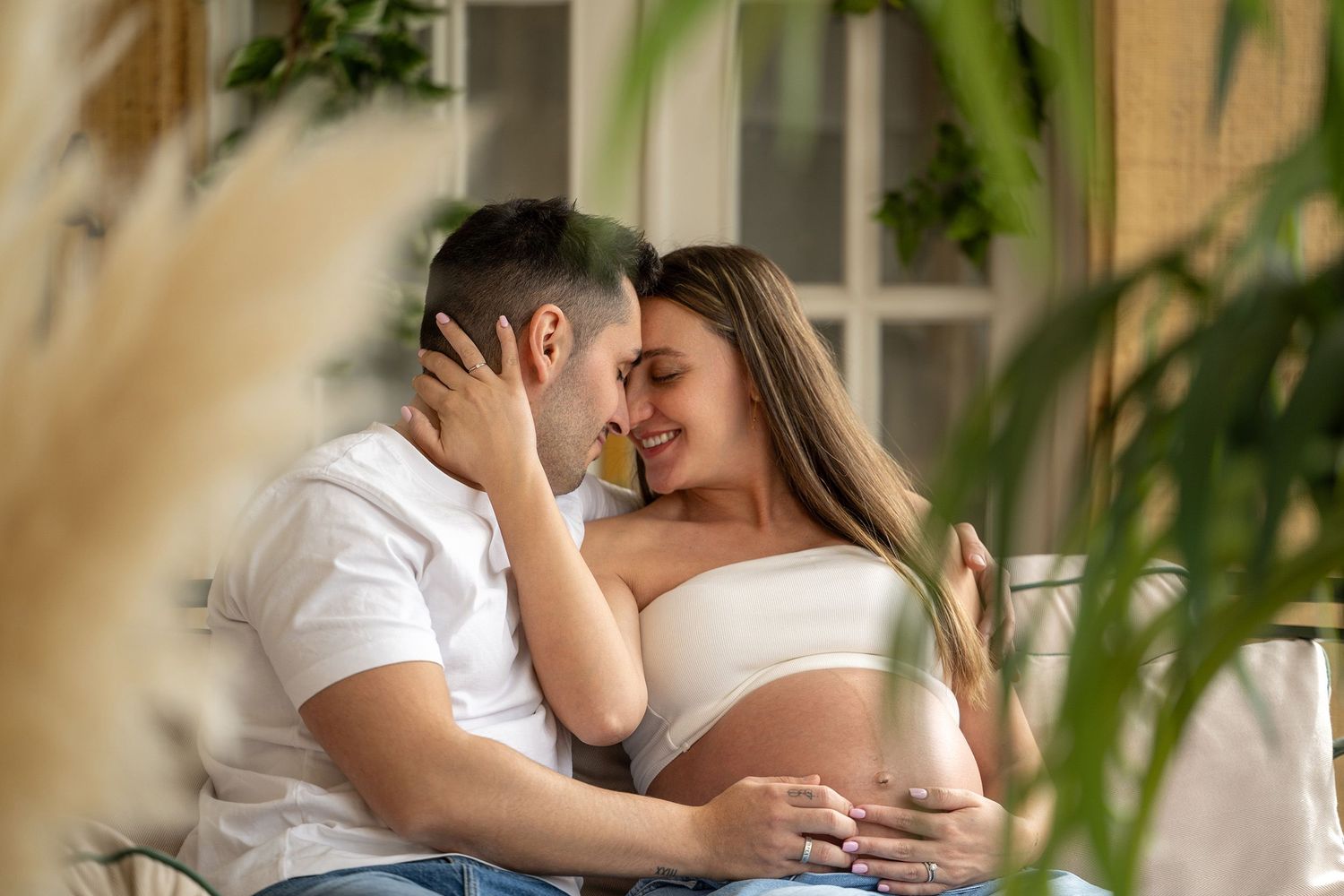 A pregnant woman in white clothing embraces her partner on a sunlit porch during a maternity photoshoot.