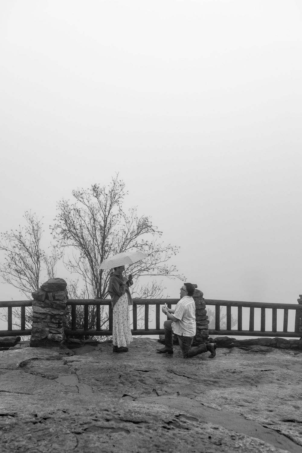 Black and white photo of man proposing on foggy stone overlook to woman.