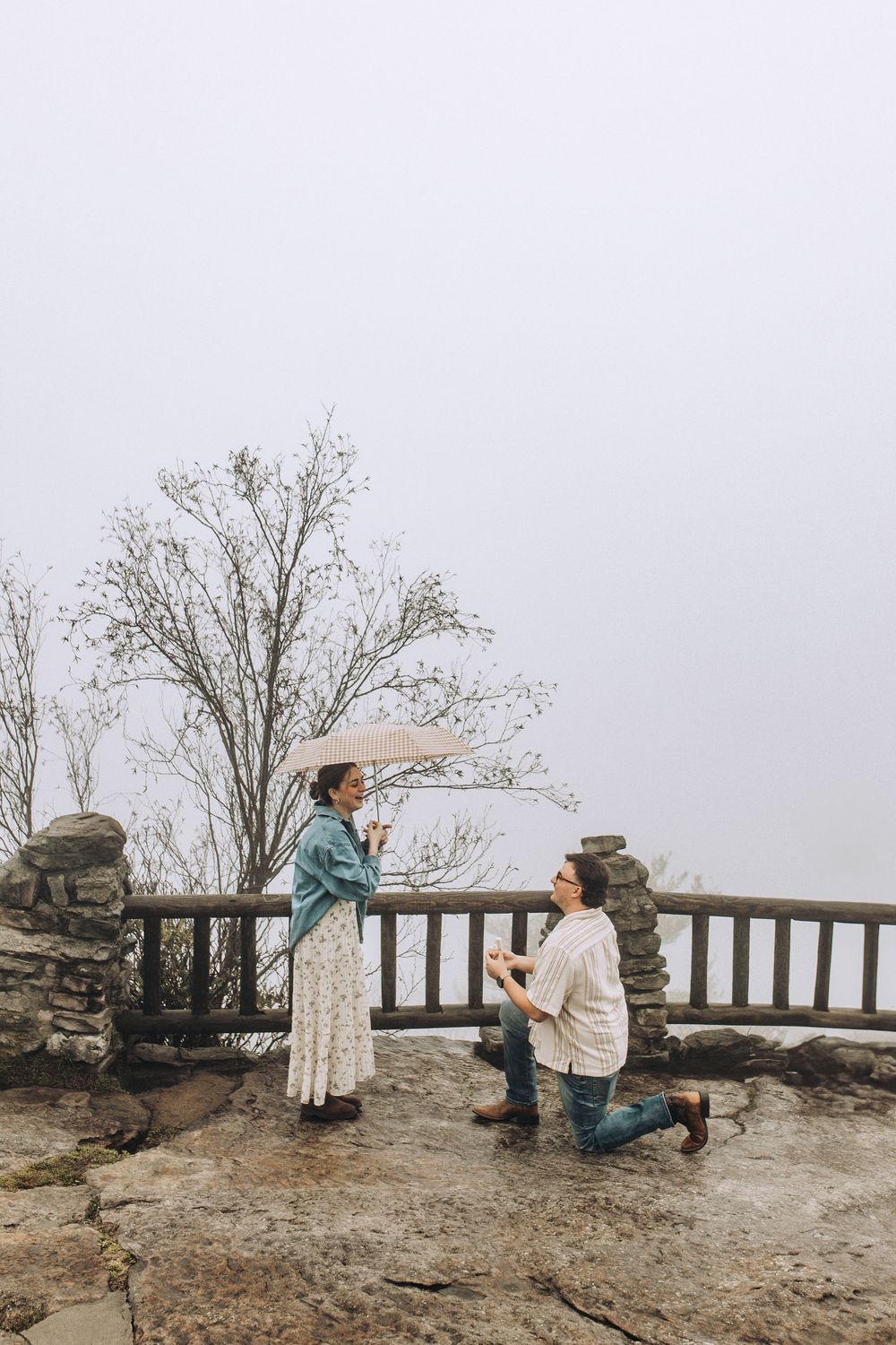 Man on one knee proposing to woman with umbrella at a foggy mountain overlook with stone fence.