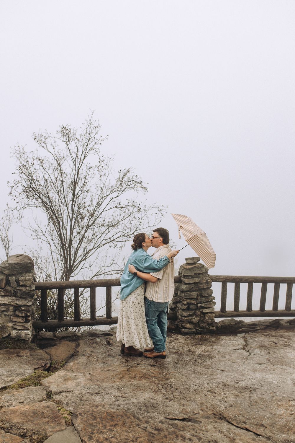 Newly engaged couple sharing a kiss under umbrella at a misty mountain overlook with stone pillars.