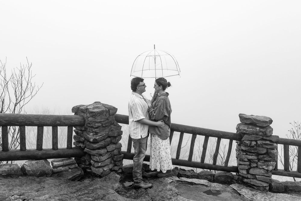 Black and white photo of a couple standing under an umbrella on a misty overlook.