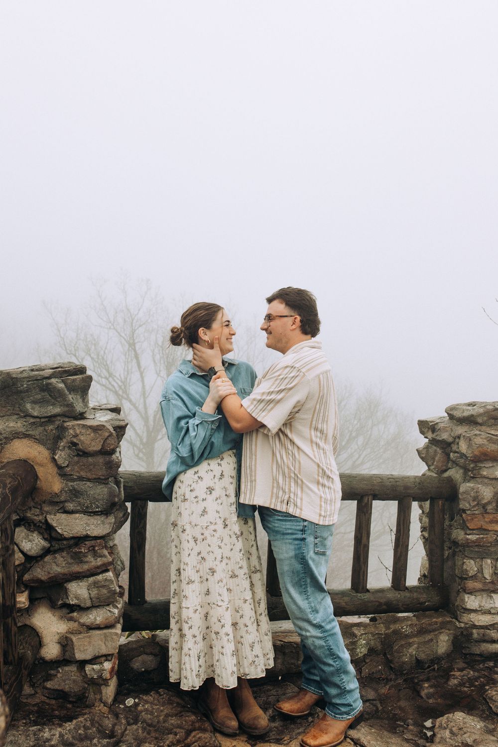 Romantic couple holding hands and gazing at each other near a stone fence in the fog.