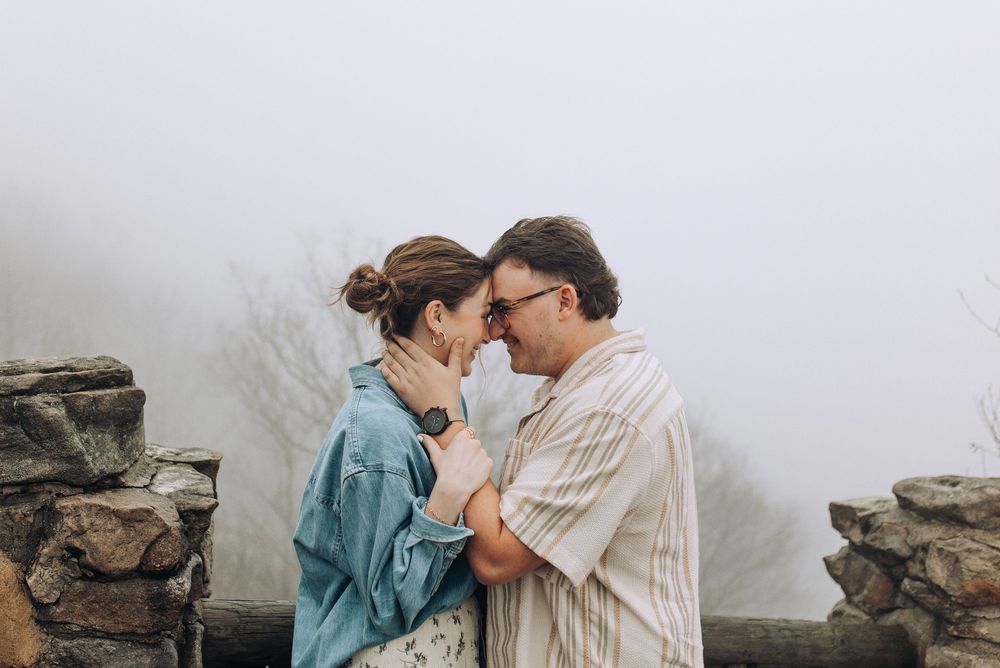 A couple shares a romantic moment, touching foreheads in a misty outdoor setting with stone walls.