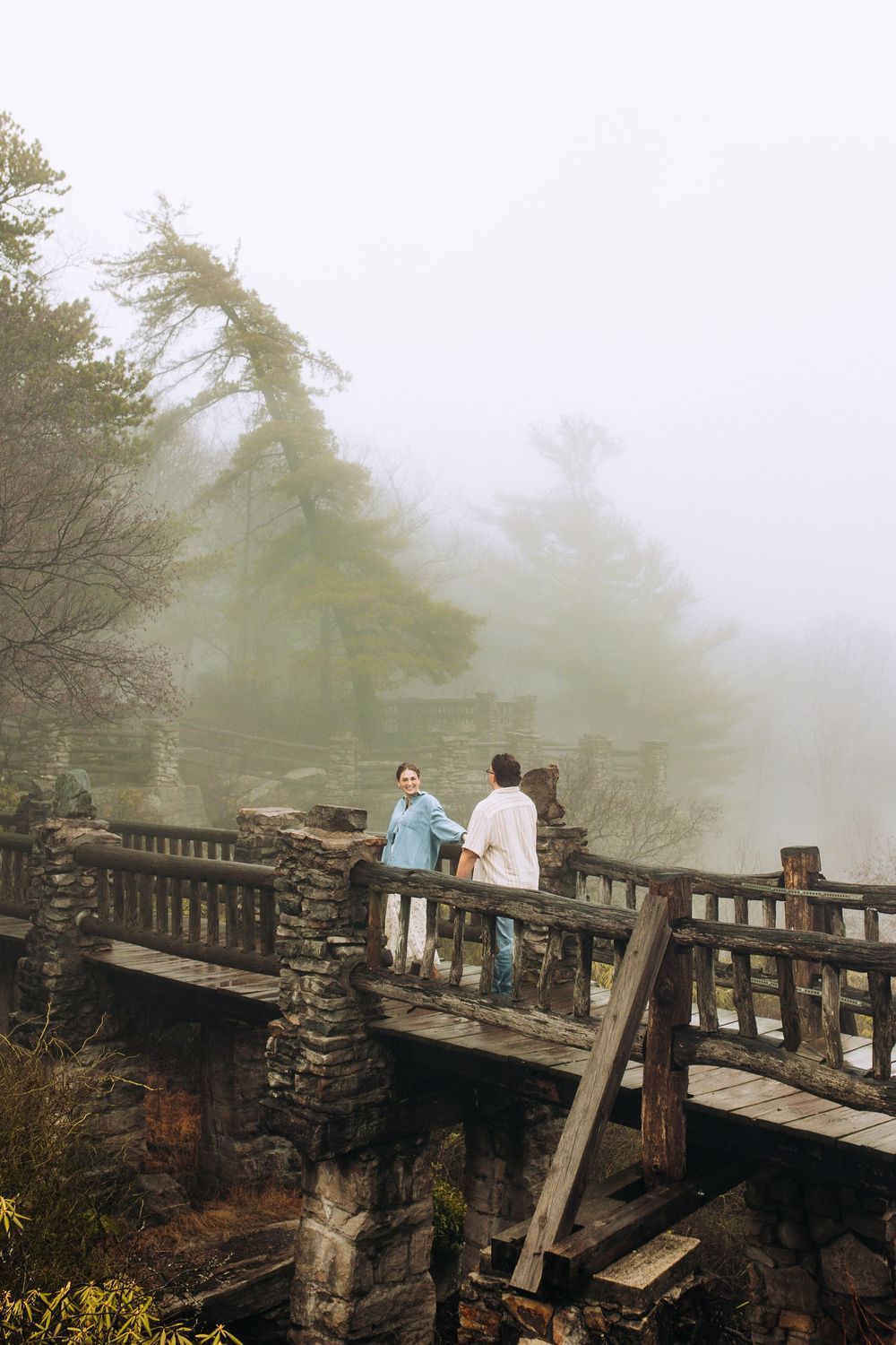 Two people sharing a moment on a stone and wood bridge in a misty woodland setting.