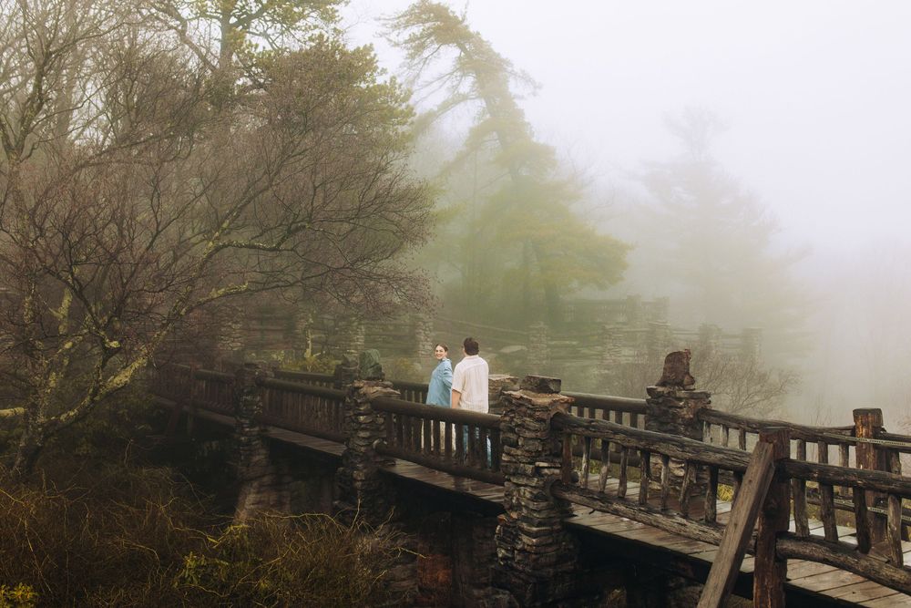 Couple strolling together on a rustic bridge surrounded by misty trees and soft light.