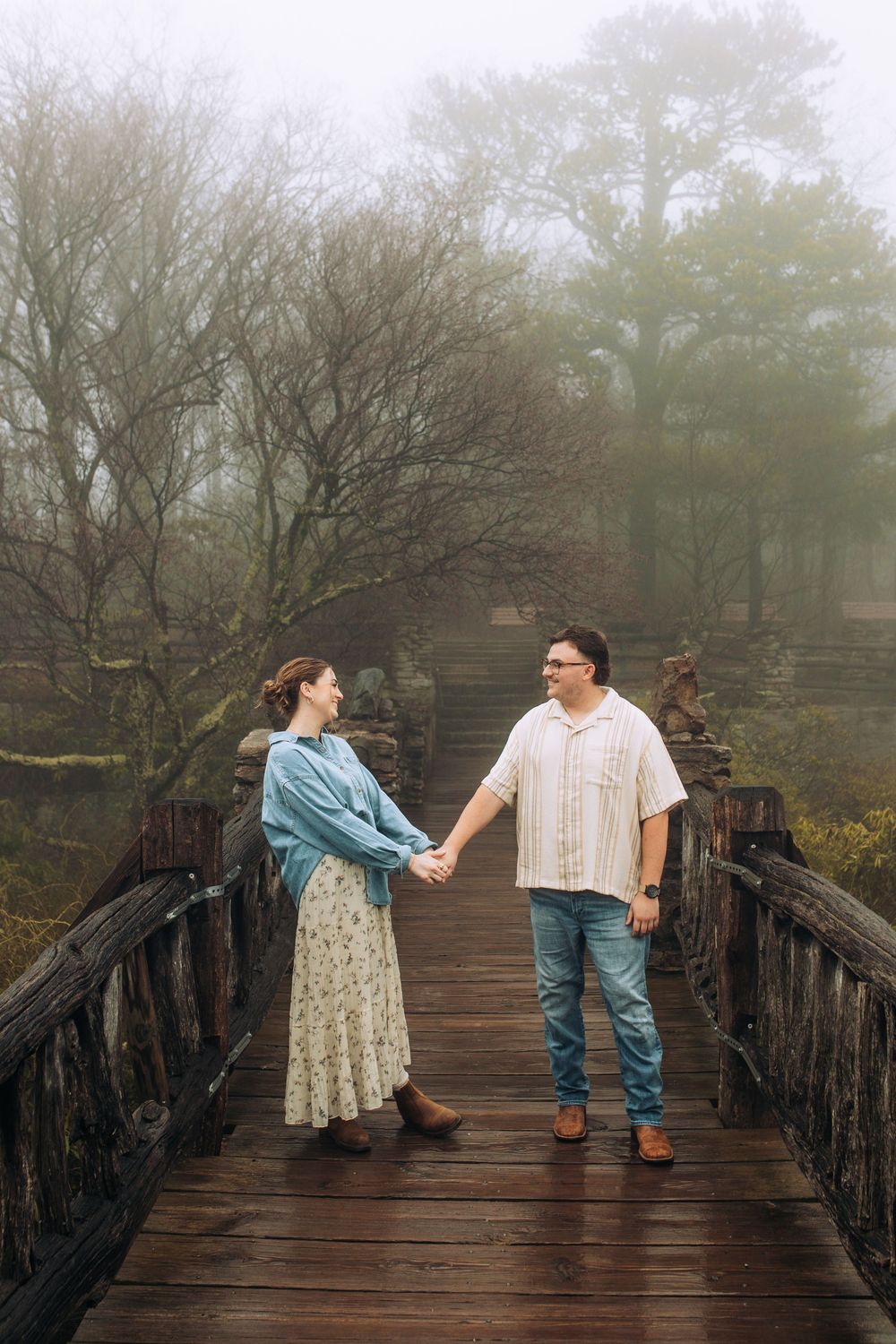 Romantic couple holding hands on a rustic wooden bridge in a foggy forest.
