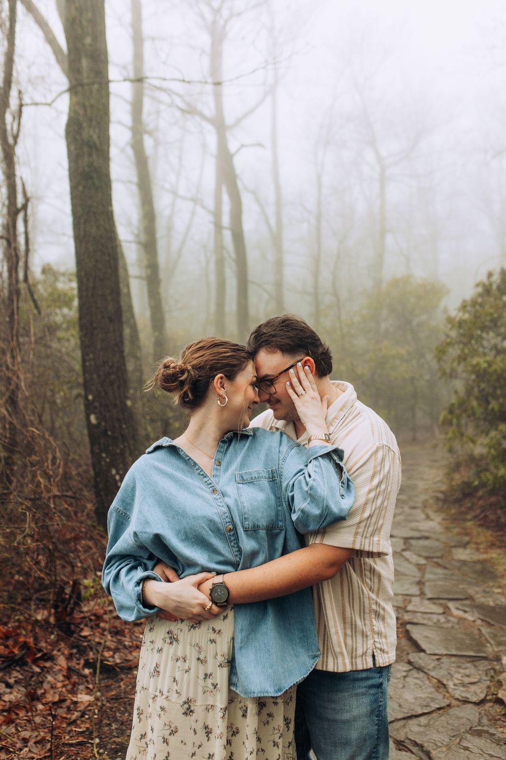 Couple sharing an intimate moment touching foreheads on a misty forest stone path.