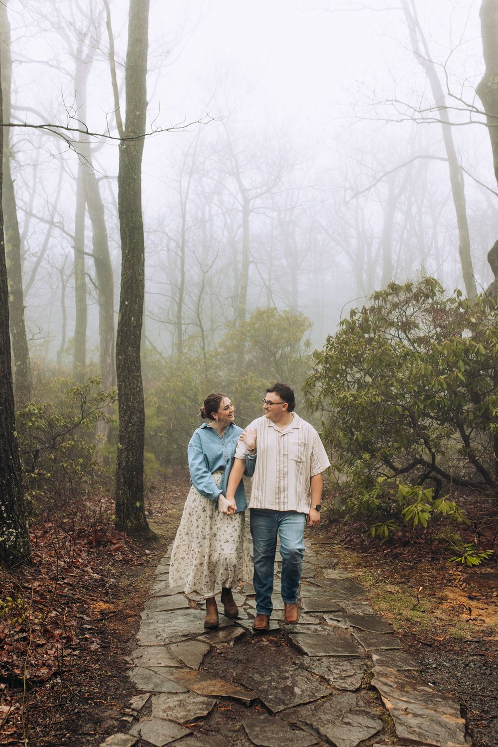 A couple walks hand in hand along a foggy stone path through a misty forest, smiling at each other.