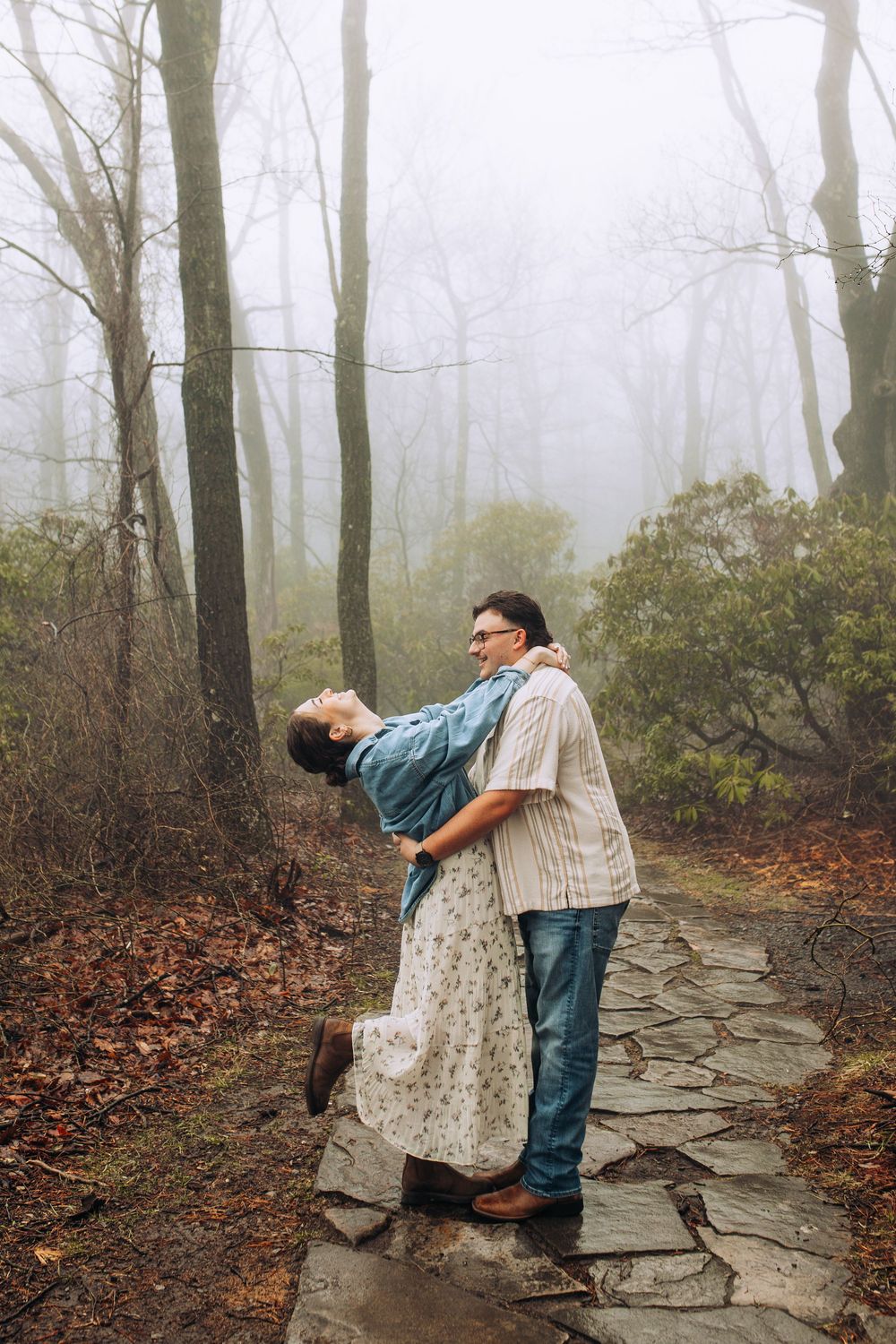 A man lifts a woman on a foggy stone forest path as she leans back joyfully in his arms.