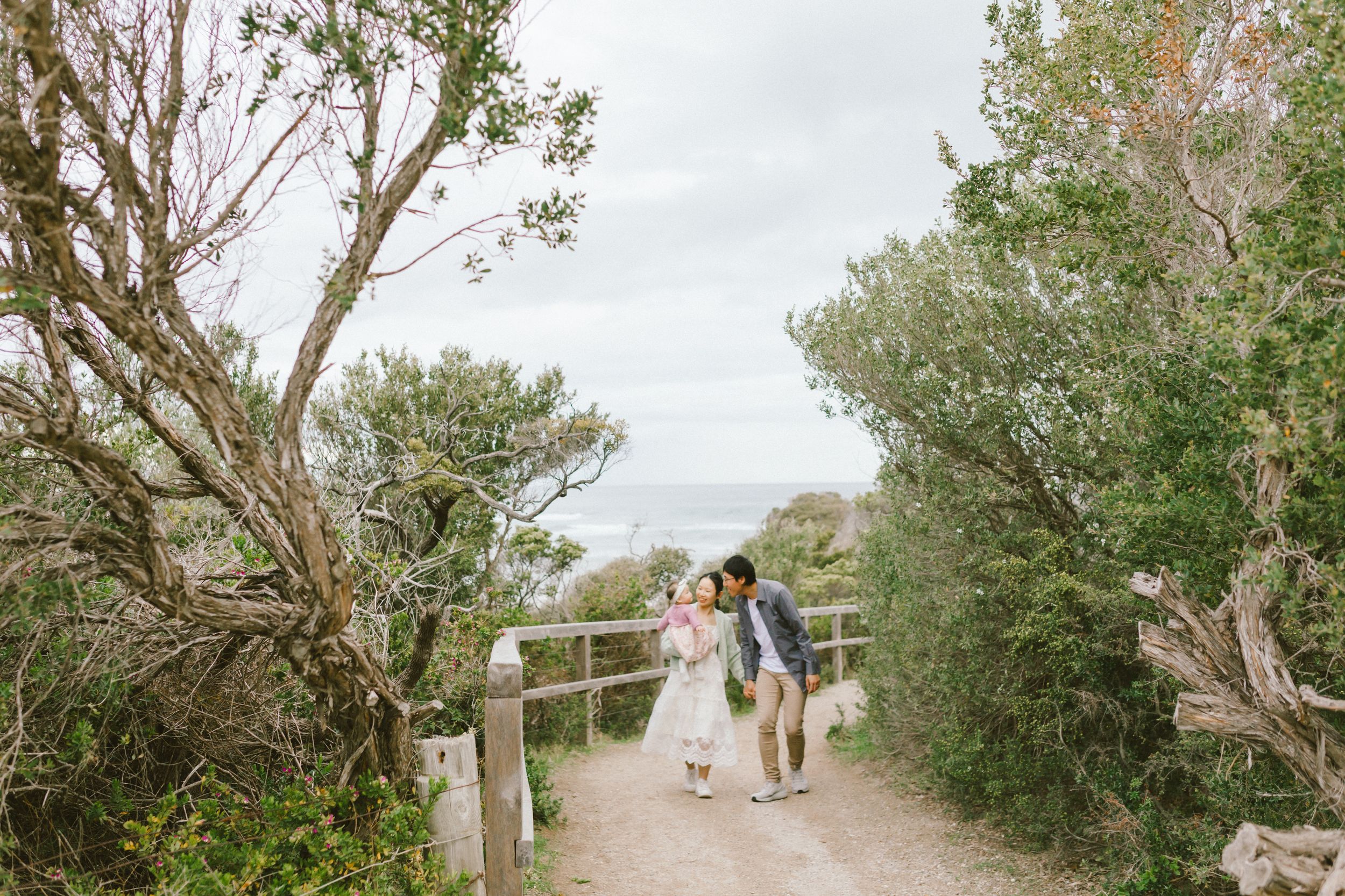 A couple walks along a scenic coastal path surrounded by lush green trees and vegetation on a cloudy day.