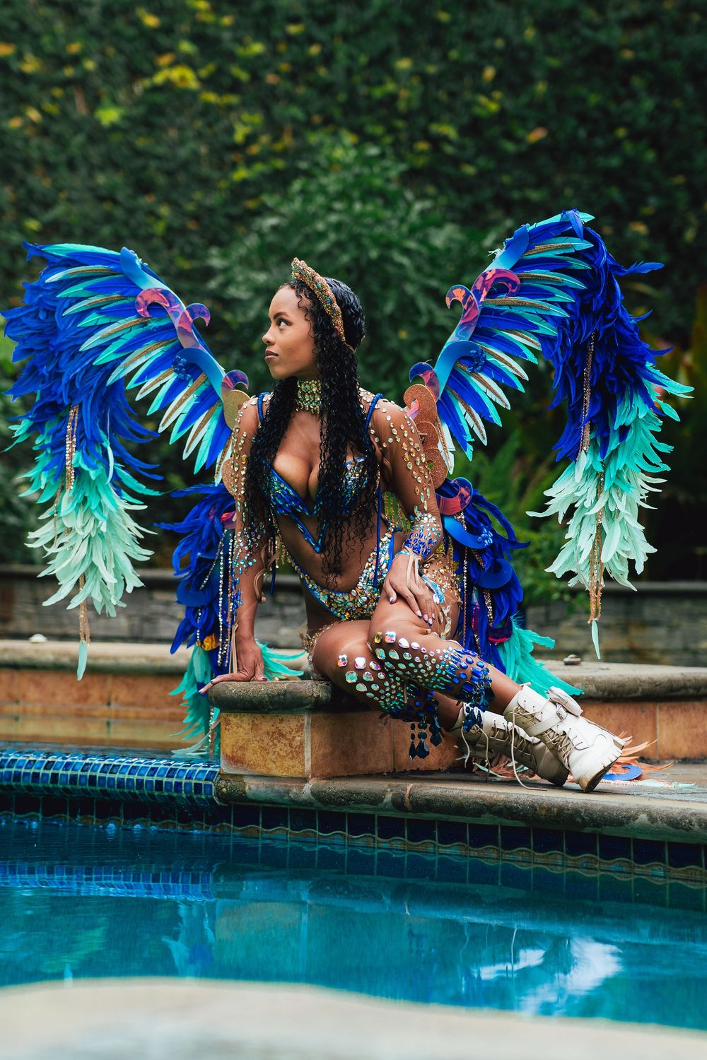 Carnival masquerader posing in a vibrant blue feathered costume beside a pool.