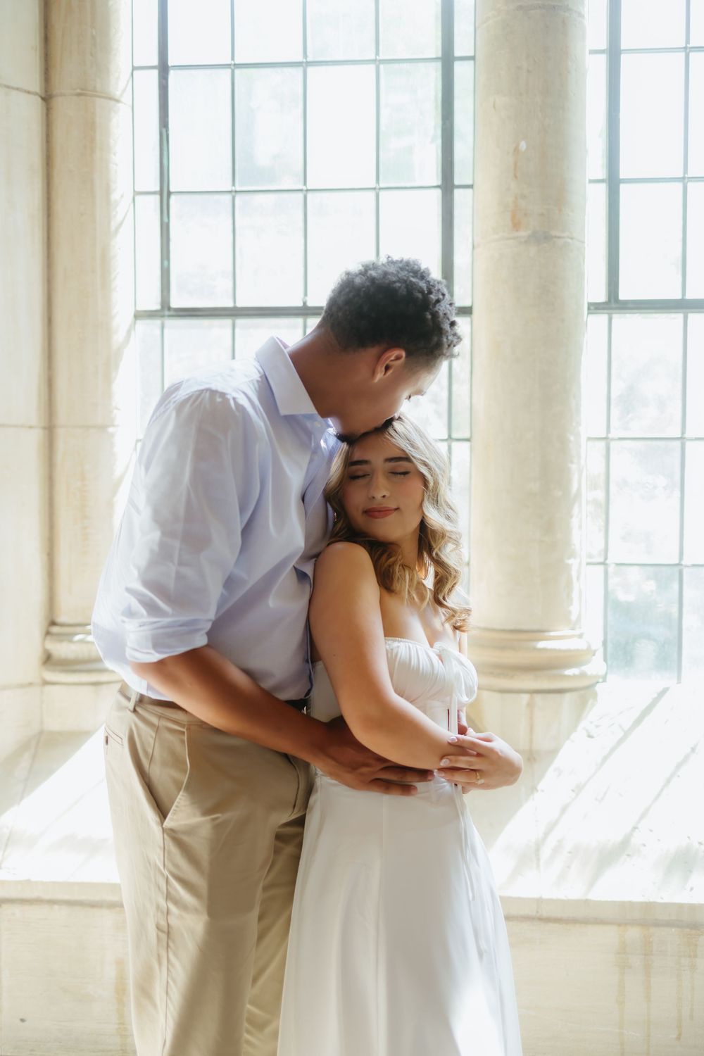 A romantic couple in white attire embraces in a bright sunlit room with elegant columns and windows.