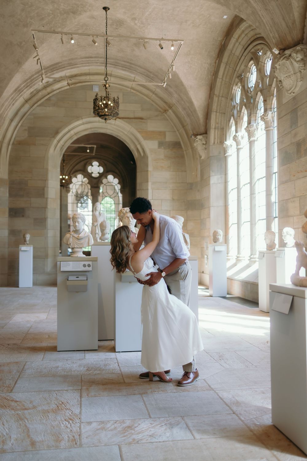 Romantic couple sharing an intimate moment in a grand stone archway corridor with gothic windows and natural light.