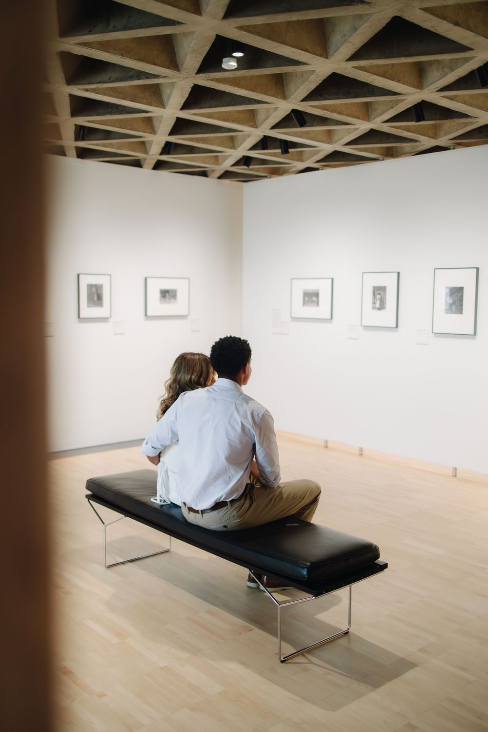 Two gallery visitors sit on a modern bench while viewing framed artwork displayed on white walls in a minimalist exhibition space.