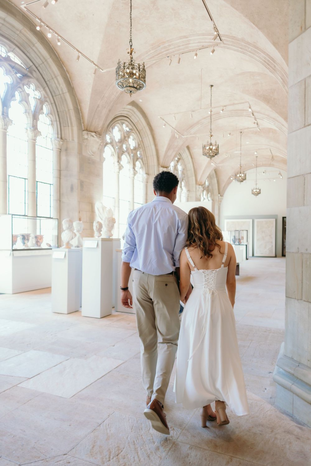 A couple walks through an elegant Gothic-style hallway with vaulted ceilings, arched windows and hanging chandeliers.