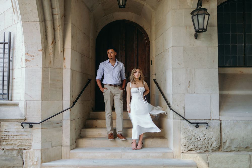 Two people walk through a stone archway corridor with antique lanterns hanging from the ceiling.