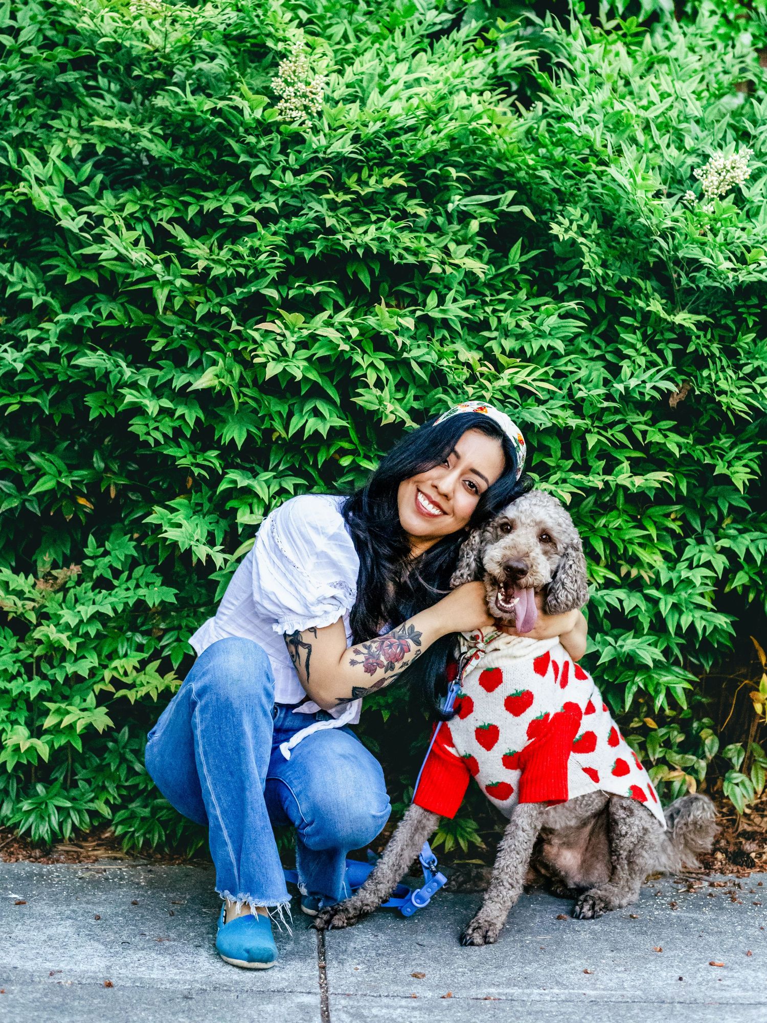 A woman in a white shirt & jeans crouches beside her dog in a strawberry-patterned outfit, smiling in front of green bushes outdoors.