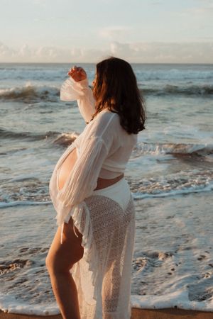 A pregnant person in white lace stands on a beach at sunset.