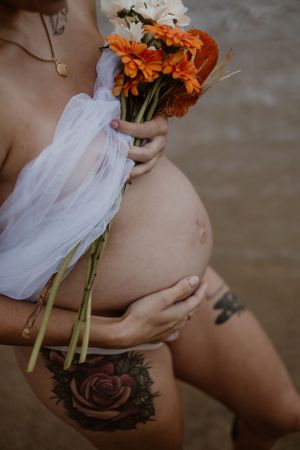 A pregnant person holds orange flowers while showing a rose tattoo in an artistic maternity photo.