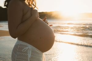 A silhouetted pregnant figure stands on beach at golden hour sunset.