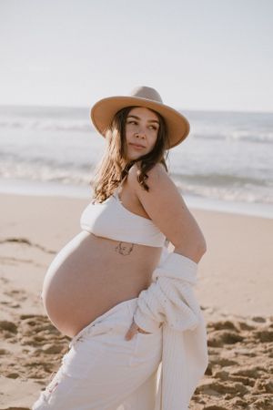 Maternity photoshoot on beach featuring white dress and wide-brim hat.