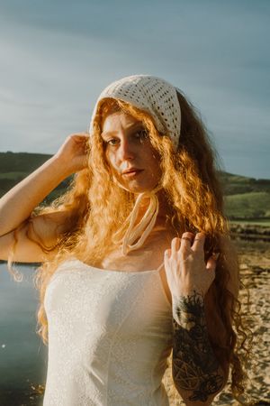 Portrait at golden hour wearing white dress and knit beanie by water.