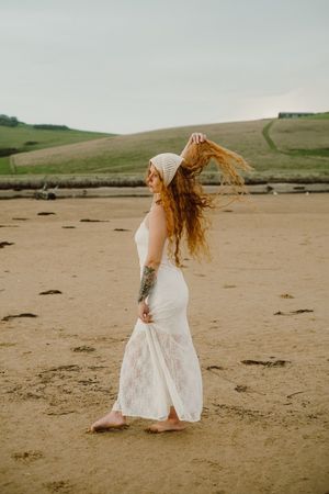 White dress flowing in breeze on sandy beach with green hills beyond.