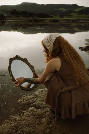 Figure in brown dress holding antique mirror by tranquil lake.