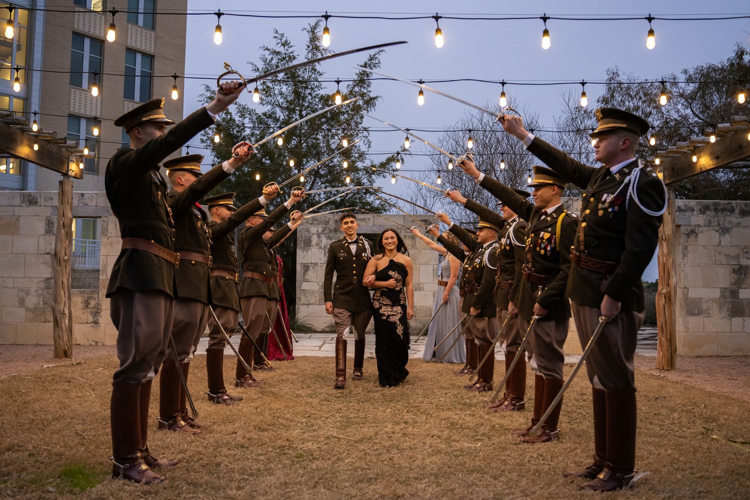 Military officers in dress uniforms form a ceremonial arch with sabers at an outdoor evening celebration with string lights.