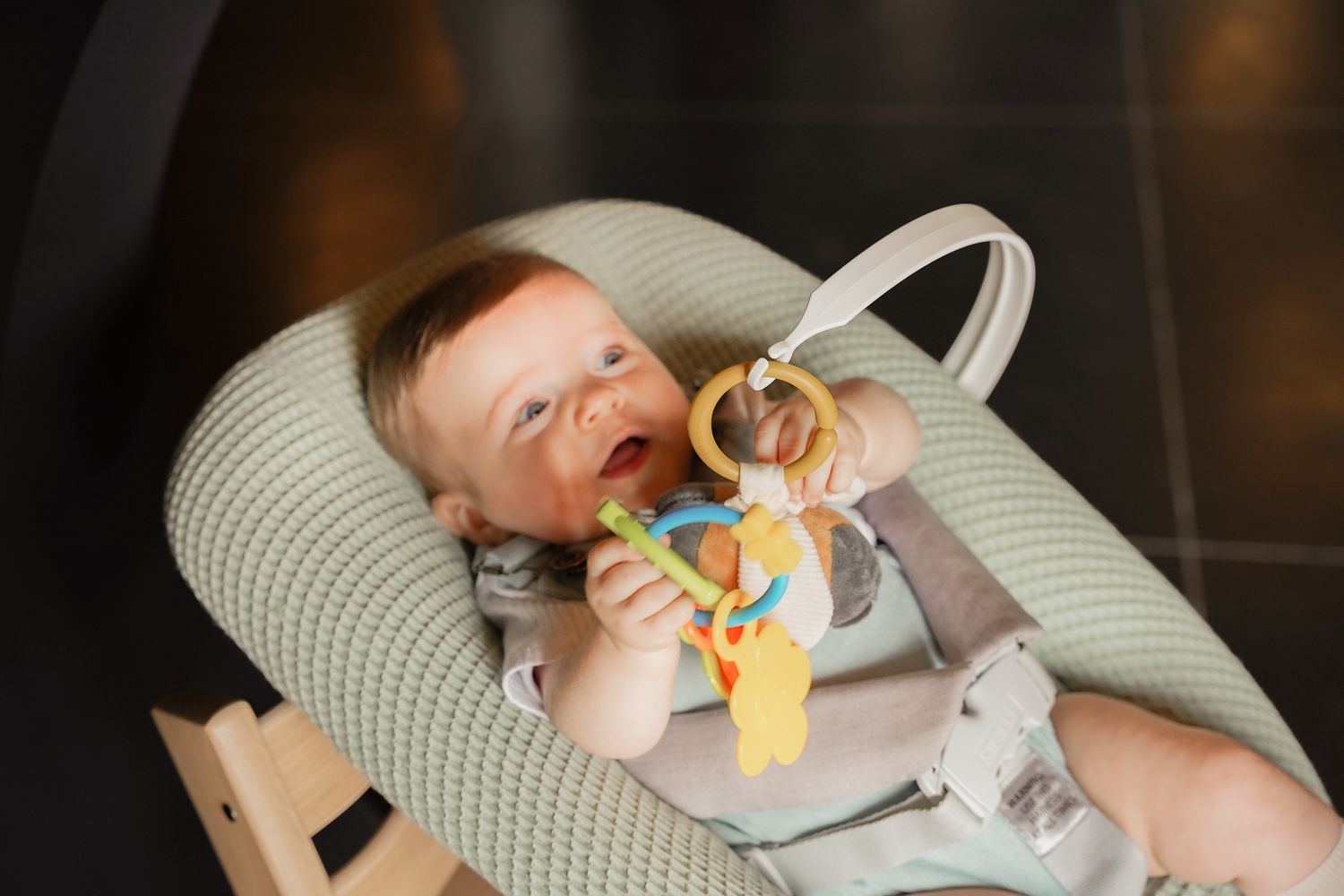 Baby lying in a grey bouncer seat playing with a colorful toy rattle.