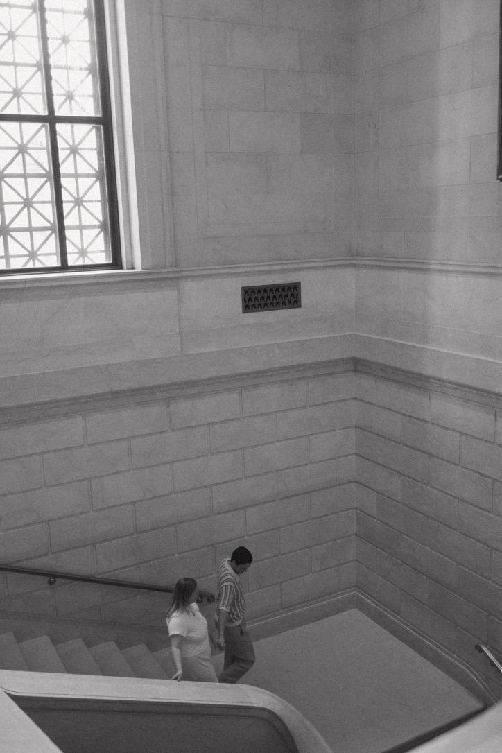 Black and white photo of a couple descending museum stairs near a large arched window.