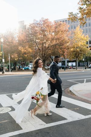 Bride in white cape gown and groom crossing a street in autumn city setting.