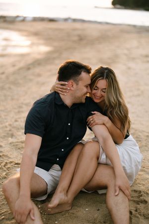 Happy couple sitting barefoot on sandy beach at sunset, embracing and smiling together.