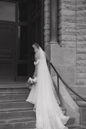 Black and white photo of bride in flowing veil ascending stone church steps with bouquet.