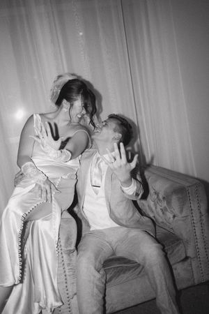 Black and white photo of playful newlyweds sitting together on a sofa at reception.