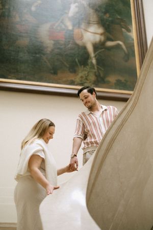 Happy couple holding hands on museum staircase beneath a classical equestrian painting.