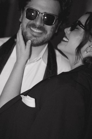 Black and white photo of a smiling couple in sunglasses, woman touching mans face at a formal event.