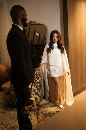 Bride in white cape top and gold skirt smiling at groom in elegant interior.
