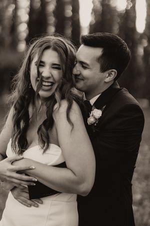 Black and white photo of laughing bride in strapless gown embraced by groom in tuxedo in forest setting.