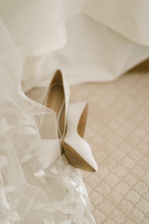 Elegant white bridal heels rest on patterned carpet beside flowing wedding veil and dress hem.