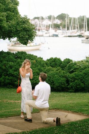 Man proposes on one knee to surprised woman covering her face near a scenic marina with sailboats.